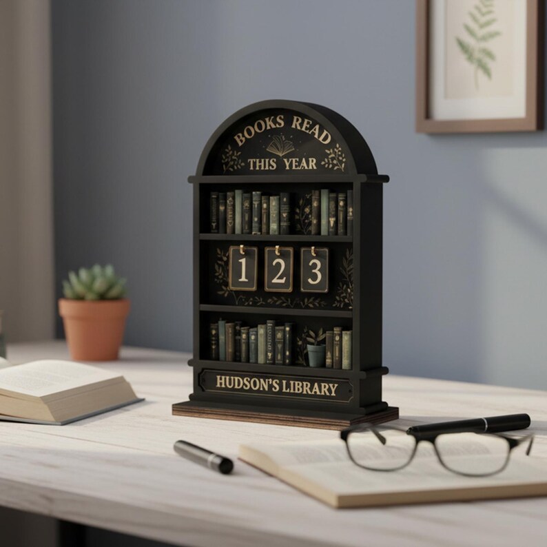May include: A black wooden bookshelf with the words "BOOKS READ THIS YEAR" and "HUDSON'S LIBRARY" in gold lettering. The bookshelf has three shelves filled with miniature books and a counter with the numbers "123".