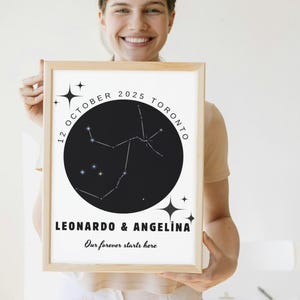May include: A framed print featuring a black circle with a constellation design, the date "12 OCTOBER 2025 TORONTO," and the names "LEONARDO & ANGELINA." The text "Our forever starts here" is also included. The frame is made of light-colored wood.