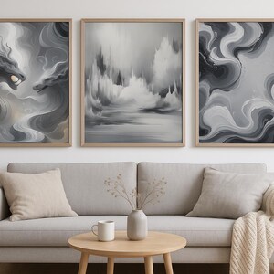 Puede incluir: Tres impresiones artísticas abstractas enmarcadas en tonos grises y blancos. La obra de arte presenta patrones arremolinados, un paisaje y una criatura parecida a un dragón. Las impresiones se exhiben sobre un sofá gris con cojines beige.