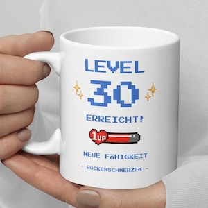 Könnte beinhalten: Weißer Keramikbecher mit dem Text "LEVEL 30 ERREICHT!" in blauer Pixel-Schrift. Der Becher zeigt eine pixelige "30" und eine "1up" Gesundheitsleiste. Zusätzlicher Text lautet "NEUE FÄHIGKEIT - RÜCKENSCHMERZEN -."