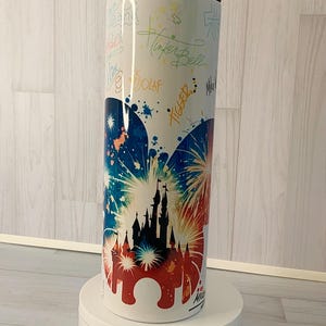 May include: White travel mug with a black lid, featuring a colorful design of a castle silhouette with fireworks in red, white, and blue. The mug is adorned with character signatures, including "Tinker Bell" and "Olaf."