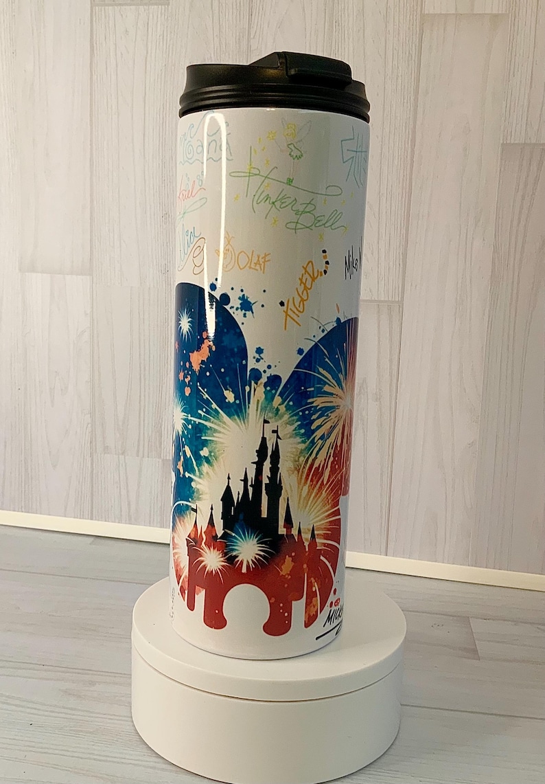 May include: White travel mug with a black lid, featuring a colorful design of a castle silhouette with fireworks in red, white, and blue. The mug is adorned with character signatures, including "Tinker Bell" and "Olaf."