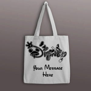 以下が含まれることがあります： 黒いディズニーのロゴと「Your Message Here」のテキストが入った白いトートバッグ。バッグには、ミッキーマウスの手袋をした手、コック帽、蝶の白黒グラフィックが描かれています。
