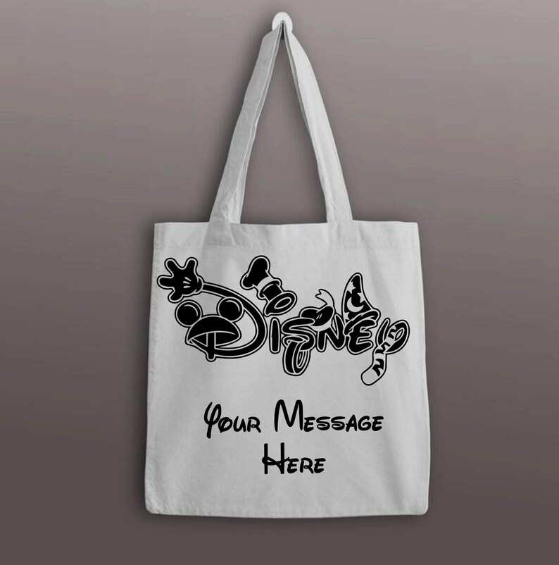 以下が含まれることがあります： 黒いディズニーのロゴと「Your Message Here」のテキストが入った白いトートバッグ。バッグには、ミッキーマウスの手袋をした手、コック帽、蝶の白黒グラフィックが描かれています。