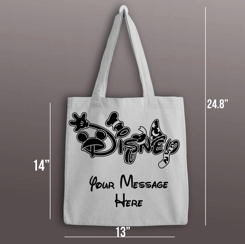 以下が含まれることがあります： 黒いディズニーのロゴと「Your Message Here」の文字が入った白いキャンバスのトートバッグ。バッグの高さは35.6cm、幅は33cm、ハンドルの上部からバッグの底部までは63cmです。