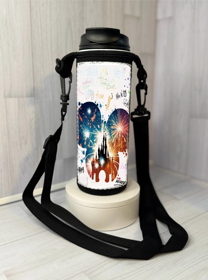 May include: White water bottle with a black lid and strap. The bottle features a colourful design of fireworks and a castle silhouette within a Mickey Mouse head shape. The design is accented with colourful signatures. The bottle is on a white stand.