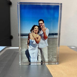 Könnte beinhalten: Ein klarer Acryl-Fotoblock, der eine Strandszene mit einem Paar zeigt. Das Paar lächelt und zeigt seine Ringe. Der Hintergrund zeigt einen blauen Himmel und das Meer. Das Foto ist in einem rechteckigen Block eingeschlossen.