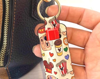 Könnte beinhalten: Ein cremefarbener Schlüsselanhänger mit einem roten Lippenbalsamhalter. Der Schlüsselanhänger zeigt ein Muster von Mickey Mouse, Popcorn, Kaffeetassen, Kameras, Herzen und Donuts. Ein goldfarbener Schlüsselring ist oben befestigt.