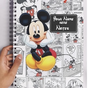 May include: A spiral-bound notebook featuring a large image of Mickey Mouse with his hands under his chin. The background is a comic strip design with various Mickey Mouse illustrations. The notebook has the text "Your Name Here" and "Notes" on a chalkboard-style label.
