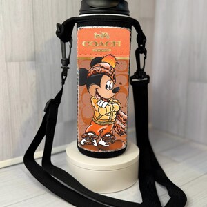 K&ouml;nnte beinhalten: Eine schwarz-orangefarbene Wasserflasche mit einem schwarzen Riemen. Die Flasche zeigt eine Mickey Mouse-Grafik und das Coach-Logo. Die Flasche steht auf einem wei&szlig;en St&auml;nder.