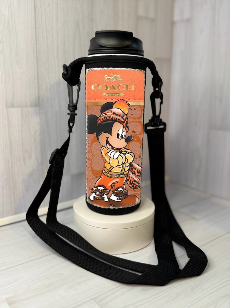 K&ouml;nnte beinhalten: Eine schwarz-orangefarbene Wasserflasche mit einem schwarzen Riemen. Die Flasche zeigt eine Mickey Mouse-Grafik und das Coach-Logo. Die Flasche steht auf einem wei&szlig;en St&auml;nder.