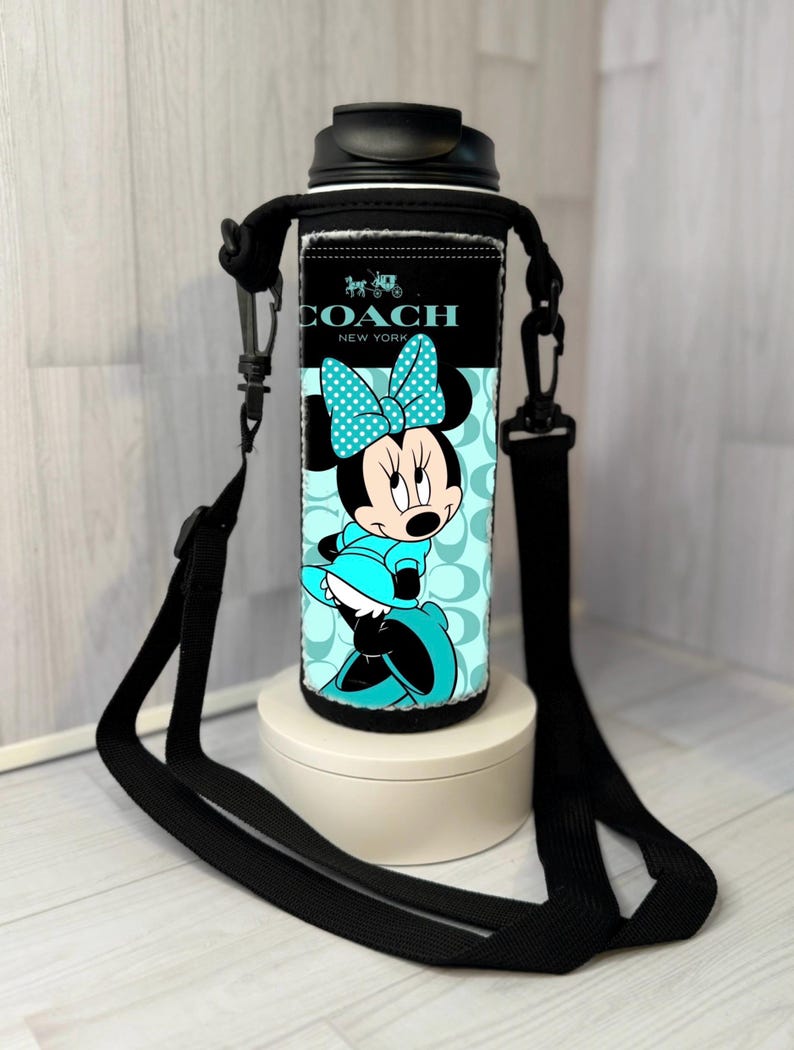 K&ouml;nnte beinhalten: Eine schwarze und t&uuml;rkisfarbene Wasserflasche mit einem Minnie Mouse-Design und dem Coach New York-Logo. Die Flasche hat einen schwarzen Deckel und einen schwarzen Trageriemen. Die Minnie Mouse-Grafik ist in T&uuml;rkis und Schwarz gehalten.