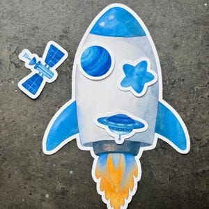 May include: Watercolor illustration of a rocket ship, a satellite, and space-themed stickers. The rocket is white and blue with a flame of orange and yellow. The satellite is blue. The stickers are on a gray background.