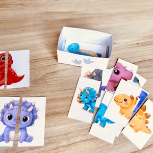 May include: A collection of colorful dinosaur-themed puzzle pieces and a small box. The puzzle pieces feature cartoon dinosaurs in red, blue, purple, and yellow. A blue dinosaur toy rests inside the box.