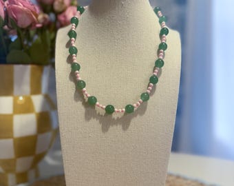 May include: A beaded necklace displayed on a mannequin. The necklace features alternating green and pink beads. The green beads are larger and more prominent, while the pink beads are smaller. The necklace is presented against a neutral background.