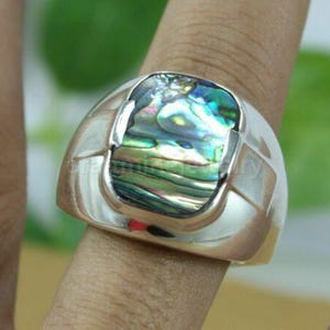 Puede incluir: Un anillo de plata con una incrustación rectangular de concha de abulón. La concha presenta colores iridiscentes verdes, azules y dorados. El anillo tiene un acabado pulido y liso, y se lleva en un dedo.