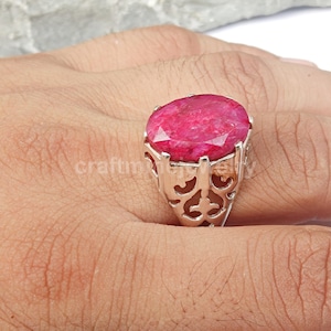 May include: A silver ring featuring a large, oval-cut, vibrant pink gemstone. The gemstone is set in a decorative, openwork silver band with an ornate design. The ring is shown on a hand.