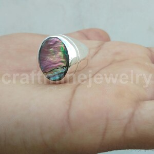 Puede incluir: Un anillo de plata con una incrustación ovalada de concha de abulón. La concha muestra colores iridiscentes de rosa, verde y azul. El anillo está colocado en una mano, con el texto "craftnejewelry" visible en el fondo.