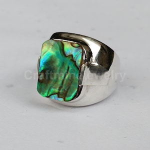 Puede incluir: Un anillo de plata con una incrustación rectangular de concha de abulón. La concha muestra tonos iridiscentes verdes, azules y dorados. El anillo tiene una banda ancha y un acabado pulido, adecuado para el uso diario.