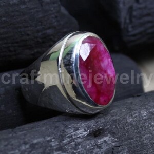 Puede incluir: Un anillo de plata con una gran gema de rubí de talla ovalada. El rubí es de color rojo intenso y está engastado en una banda de plata pulida. El anillo se muestra sobre una superficie oscura y texturizada, con el texto "craftminejewelry".
