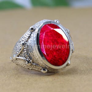 Könnte beinhalten: Ein silberner Ring mit einem großen, oval geformten, tiefroten Edelstein. Der Ring hat ein strukturiertes Band mit dekorativen Elementen. Der Edelstein ist in einer polierten Silberfassung gefasst, was einen auffälligen Kontrast erzeugt.