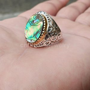 Puede incluir: Anillo de plata con una incrustación ovalada de concha de abulón. El anillo tiene un diseño decorativo en la banda y un bisel dorado alrededor de la concha. La concha de abulón muestra colores iridiscentes verdes, azules y dorados.
