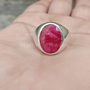 Puede incluir: Un anillo de plata con una gran gema de rubí ovalada y facetada. El rubí es de un color rojo intenso y vibrante. El anillo está engarzado en una mano, centrándose en la joya. El anillo es una pieza llamativa.