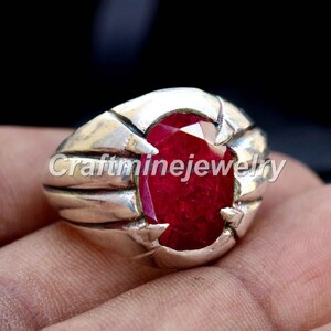 May include: A silver ring featuring a large, oval-cut ruby gemstone. The ruby is a deep red hue, set in a silver band with a textured design. The ring is shown on a hand.