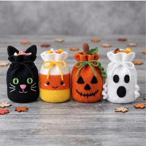 May include: Four crocheted drawstring pouches featuring Halloween themes: a black cat with green eyes, a yellow and orange candy corn, an orange pumpkin with a brown stem and green vine, and a white ghost, all filled with candy corn pieces.