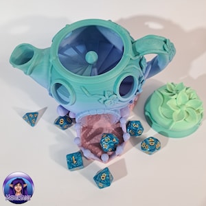 May include: A teal and blue teapot shaped dice tower with a floral design. The tower has a removable lid and a small opening at the top. There are several blue dice with gold numbers scattered around the tower.