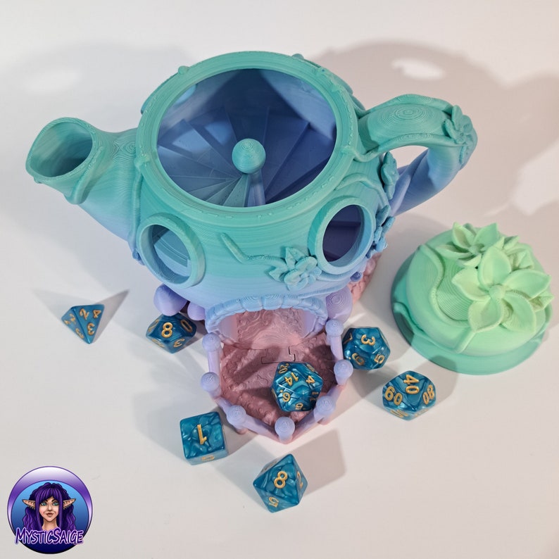 May include: A teal and blue teapot shaped dice tower with a floral design. The tower has a removable lid and a small opening at the top. There are several blue dice with gold numbers scattered around the tower.