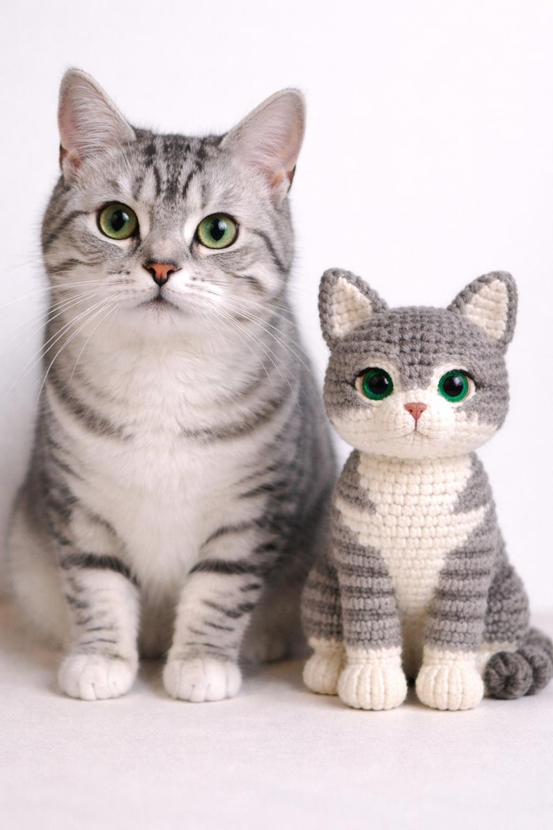 Op de afbeelding: Een grijze tabby kat zit naast een gehaakte kat. De echte kat heeft groene ogen en een grijs en wit gestreepte vacht. De gehaakte kat is grijs en wit met groene ogen en is een handgemaakt item.