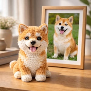 Op de afbeelding: Een gehaakte Shiba Inu hond figuur in oranje en wit, met een vrolijke uitdrukking en zijn tong zichtbaar. Een ingelijste foto van een Shiba Inu hond staat op de achtergrond. Het beeldje staat op een houten oppervlak.