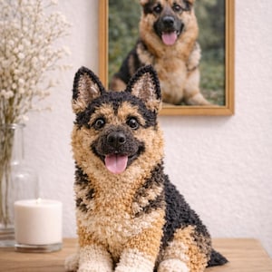Op de afbeelding: Handgemaakte pluche sculptuur van een Duitse herdershond met een glimlachende uitdrukking en een roze tong. De hond is zwart, bruin en cr&egrave;mekleurig en zit op een houten oppervlak. Een ingelijste foto van een Duitse herdershond hangt op de achtergrond.