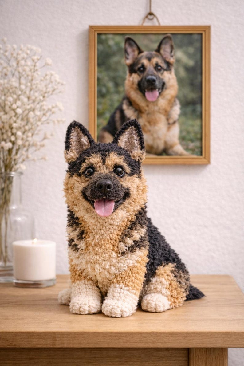 Op de afbeelding: Handgemaakte pluche sculptuur van een Duitse herdershond met een glimlachende uitdrukking en een roze tong. De hond is zwart, bruin en cr&egrave;mekleurig en zit op een houten oppervlak. Een ingelijste foto van een Duitse herdershond hangt op de achtergrond.