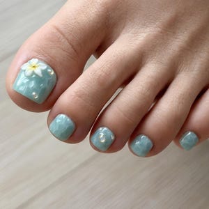 May include: Toenails painted with a glossy, light blue textured polish that mimics the appearance of water ripples. The big toe features a small, white, five-petaled flower embellishment and tiny, clear, round accents on the nail surface.