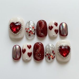 May include: A set of ten press-on nails featuring a variety of designs including cream-colored bases with large red heart-shaped rhinestones, silver and red glitter accents, deep red swirl patterns, small red hearts, and iridescent chrome finishes.