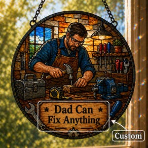 Puede incluir: Panel de vidriera redondo que representa a un artesano en un taller, martillando un clavo. El panel incluye una caja de herramientas, herramientas y el texto "Dad Can Fix Anything". El fondo muestra una ventana y una lámpara colgante.