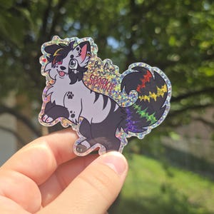 May include: A holographic sticker featuring a cartoon dog with black and white fur and a rainbow tail. The sticker has the words "Be free and Sparkle" printed on it.