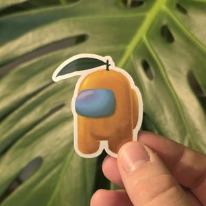 May include: A sticker featuring a yellow Among Us character designed as an orange, complete with a green leaf and stem. The character has a blue visor. The sticker is held in front of a green leafy background.