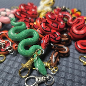 Puede incluir: Una colección de coloridos amuletos con forma de serpiente. Los amuletos son de varios colores, incluyendo rojo, verde, amarillo y marrón, con patrones intrincados. Algunos amuletos tienen cierres dorados, lo que sugiere que son llaveros o accesorios.