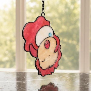 May include: Stained glass suncatcher featuring a cartoon character with red, white, and peach tones, outlined in black. A small blue gem accents the design. The suncatcher hangs from a black chain, perfect for window decor.