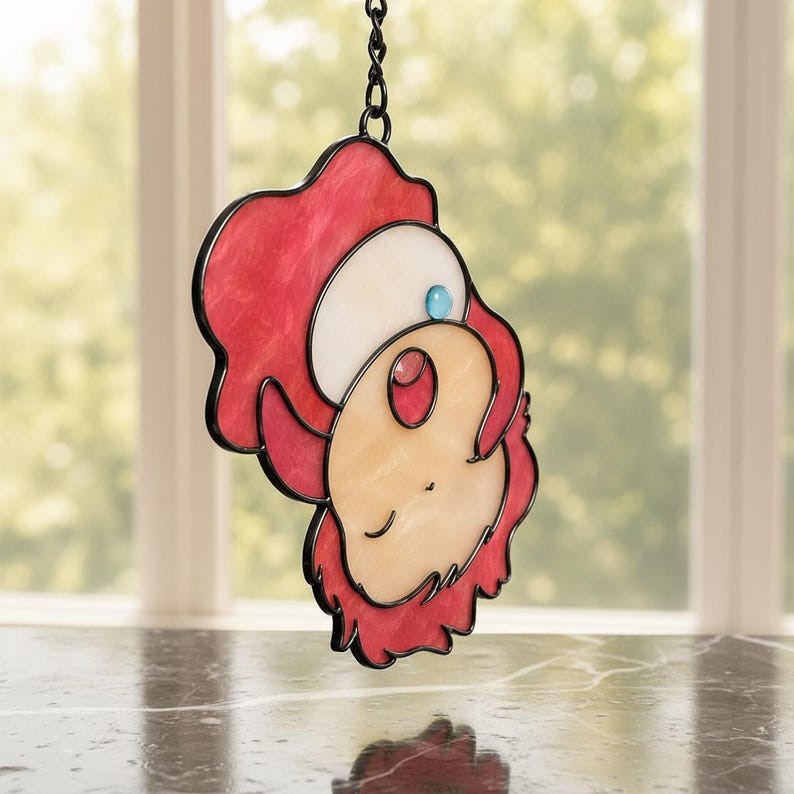 May include: Stained glass suncatcher featuring a cartoon character with red, white, and peach tones, outlined in black. A small blue gem accents the design. The suncatcher hangs from a black chain, perfect for window decor.