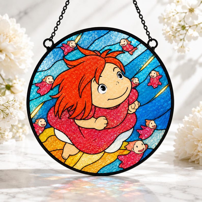 May include: A circular stained glass suncatcher featuring a central character with bright red hair and a pink dress, surrounded by small red fish figures against a blue wavy background. The piece is framed in black metal with a hanging chain attached.