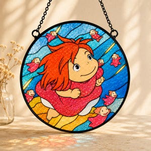 May include: A circular stained glass suncatcher featuring a central character with bright orange hair and a red dress, surrounded by small red fish figures against a blue wavy background. The piece is framed in black metal with a hanging chain.