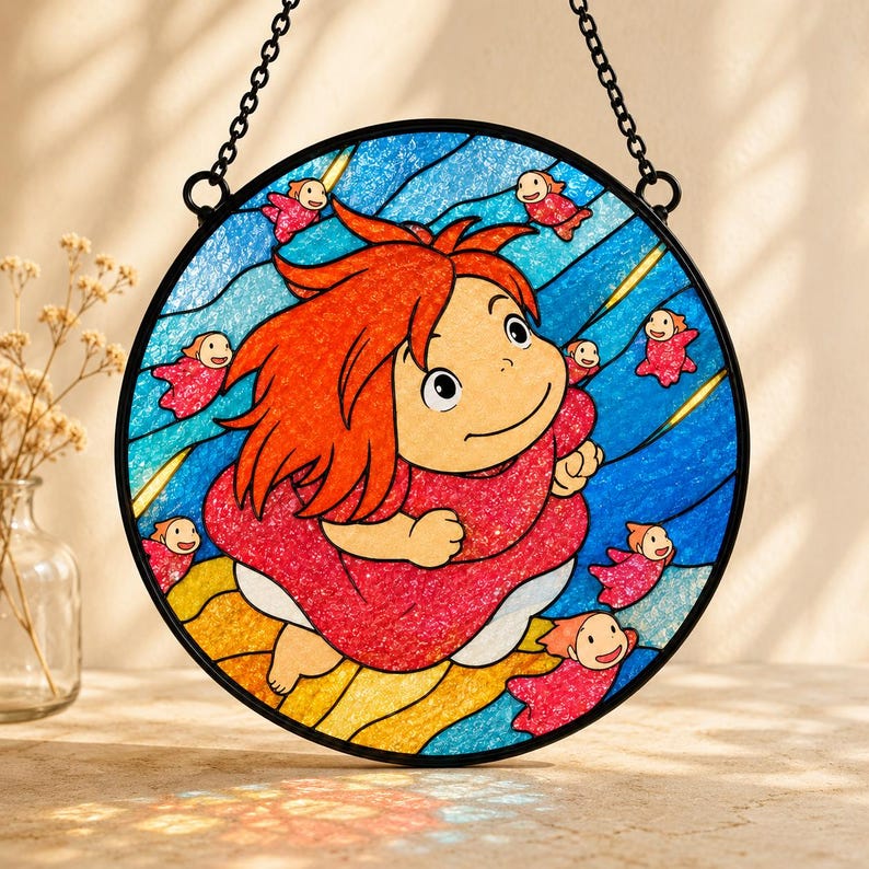 May include: A circular stained glass suncatcher featuring a central character with bright orange hair and a red dress, surrounded by small red fish figures against a blue wavy background. The piece is framed in black metal with a hanging chain.