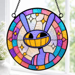 Puede incluir: Un atrapasoles circular de vidrieras que presenta un personaje morado parecido a un conejo con una amplia sonrisa amarilla. El diseño está enmarcado por un borde de segmentos de vidrio multicolor en azul, rosa y amarillo, suspendido por una cadena de metal negro.