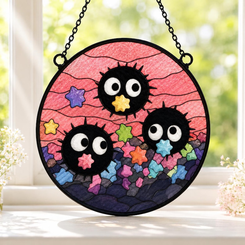May include: A circular stained glass suncatcher featuring three black, spiky, round soot sprites with large white eyes. The background is a pink sky with wavy lines, and the bottom is filled with dark blue, purple, and colorful star-shaped pieces.