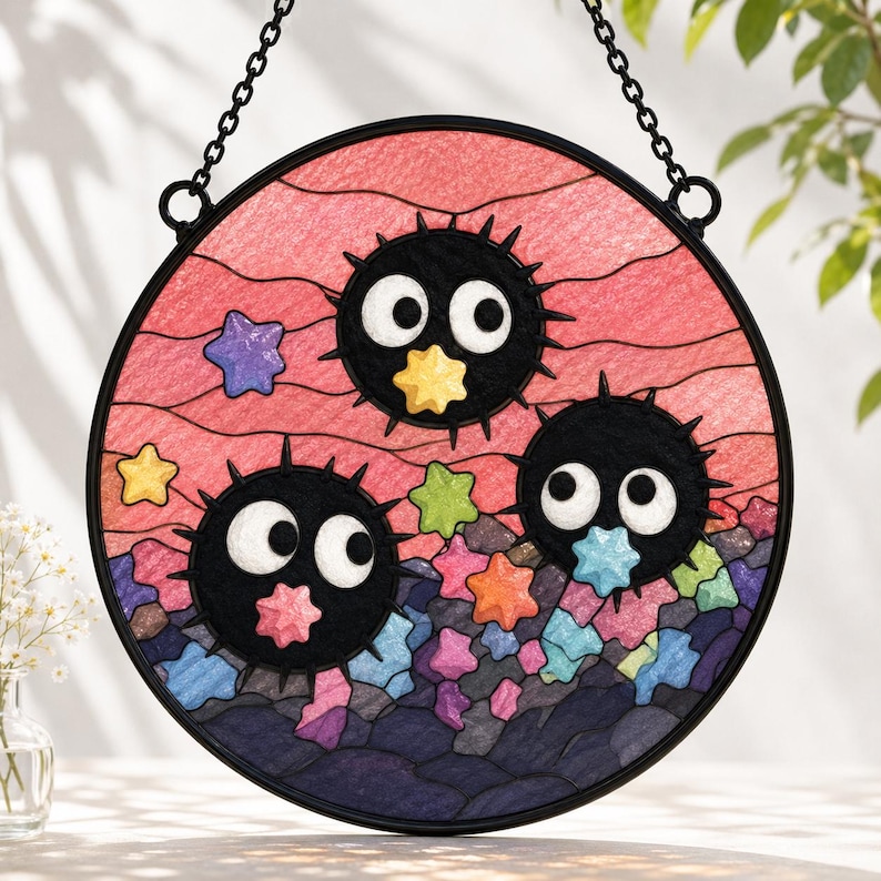May include: A circular stained glass suncatcher featuring three black spiky soot sprites with large white eyes. The background is a textured pink sky with wavy lines and a collection of colorful small stars in shades of blue, yellow, and green.