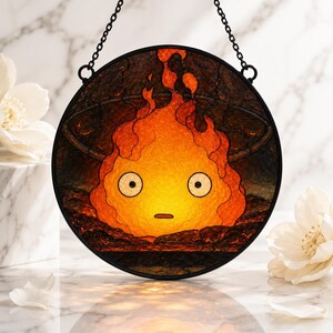 Puede incluir: Un atrapasoles circular de vidriera que presenta un personaje de llama naranja y amarillo estilizado con grandes ojos blancos y una boca peque&ntilde;a, enmarcado en metal negro con una cadena, mostrando una superficie texturizada y detalles oscuros.